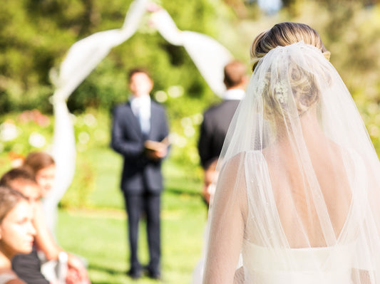 A close up photo of the back of a bride, her veil hanging down. In the background, a groom waits under a wedding arch.