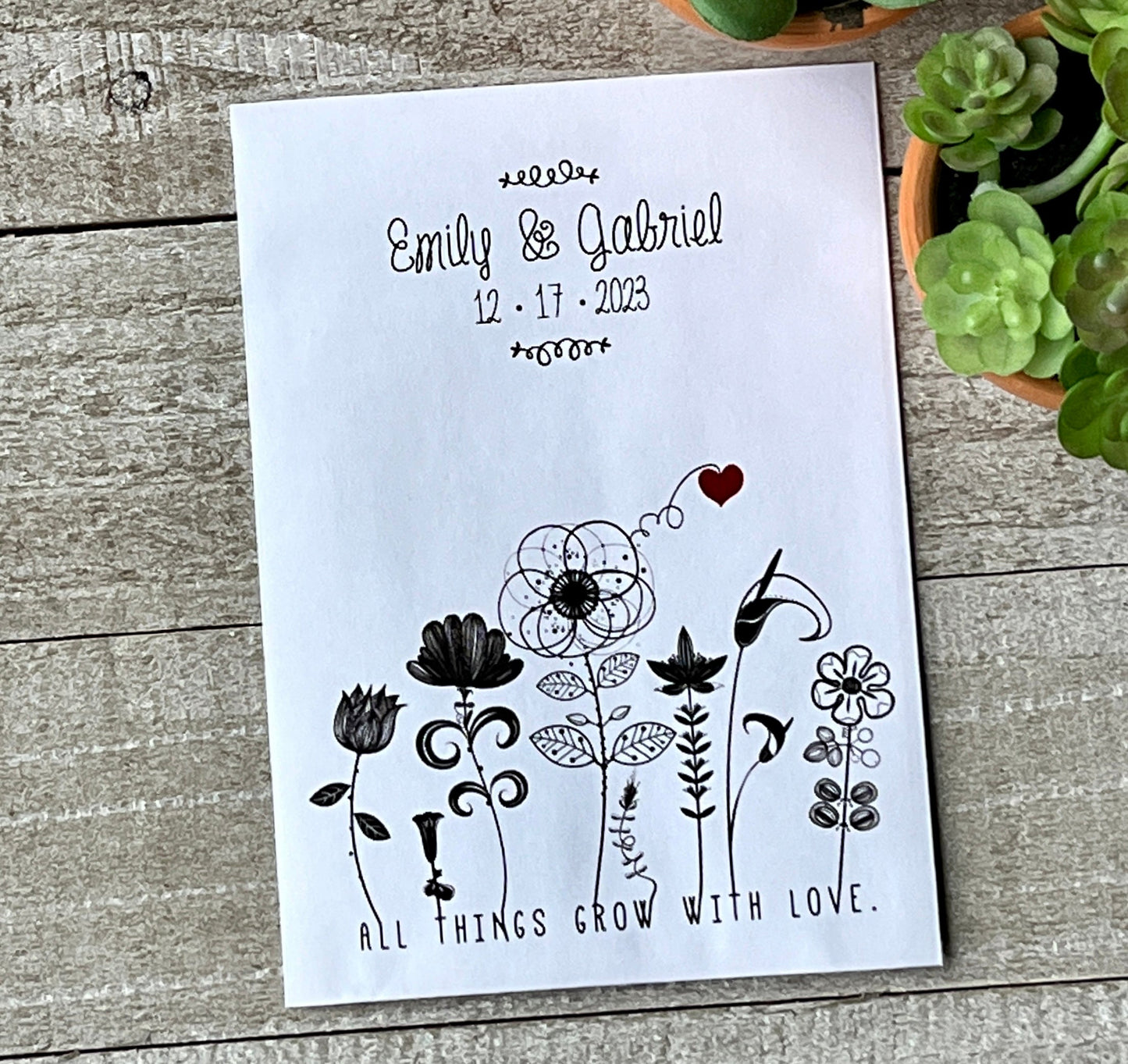 A white seed packet wedding favor with black line-drawn flowers and a red heart on the bottom edge.